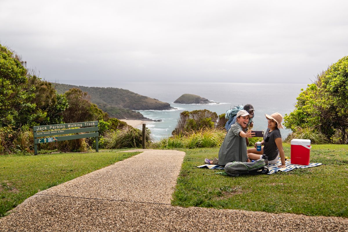Three people taking a selfie on a picnic Captain Cooks Lookout, Arakoon National Park. Photo credit: Rob Mulally / DCCEEW © Studio Bilby