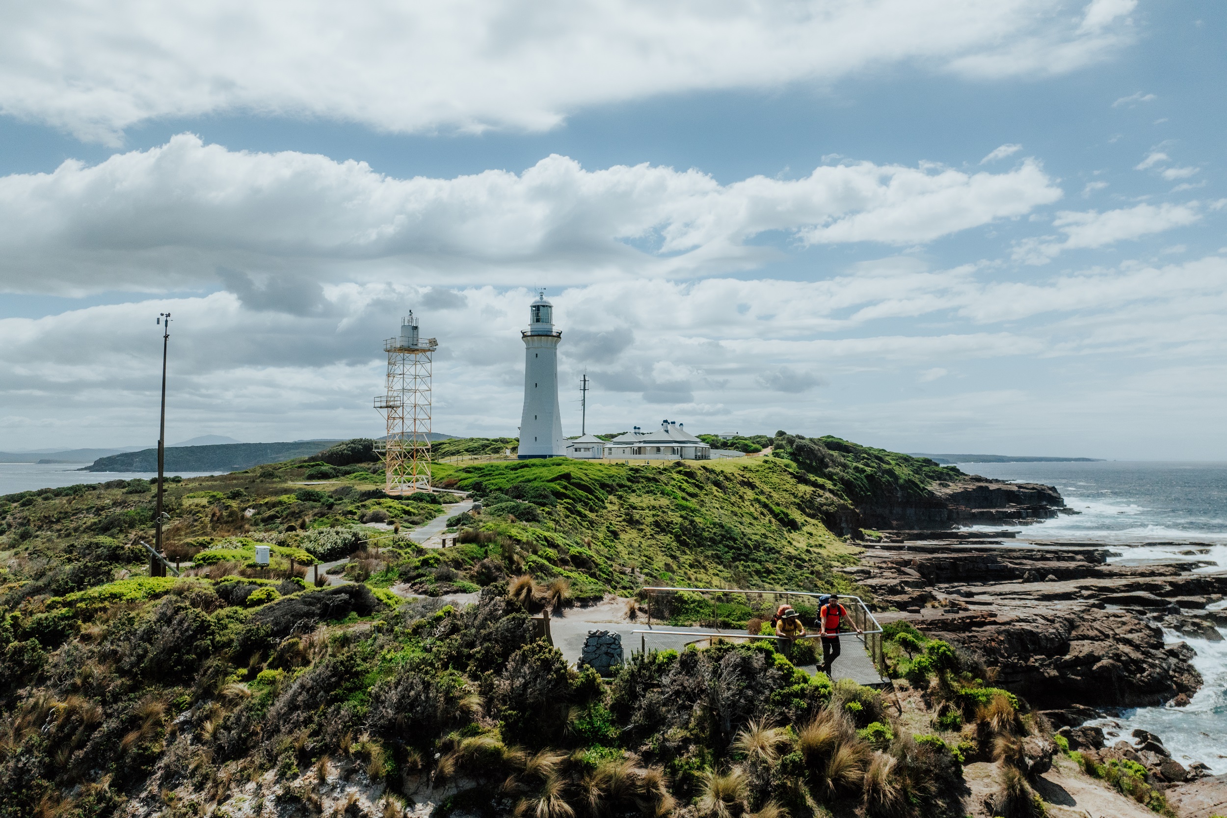 Two walkers take in veiws from the lookout at Green Cape Lighthouse on the Light to Light Walk. Photo credit: Daniel Parsons/DCCEEW © Studio Bilby