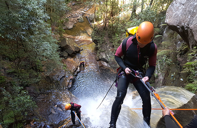 A pair of wetsuit-clad climbers use ropes to abseil past a waterfall as they descend into a steep canyon. Photo credit: James Waddell  &copy; Eagle Rock Adventures