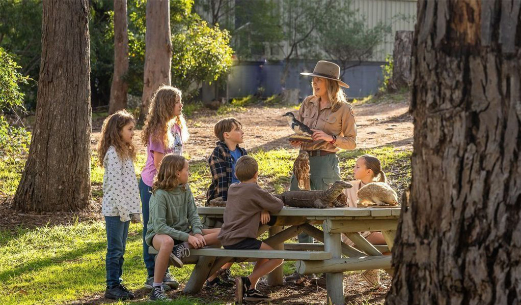 A NPWS guide talking to a kids' tour group sitting at a picnic table in a park. Credit: David Rogers &copy; DCCEEW