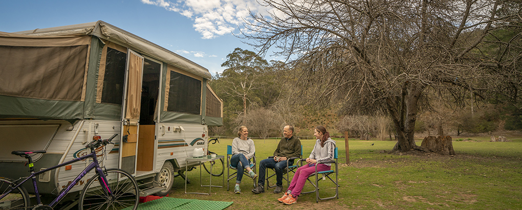 Three people sit on camp chairs beside a caravan at Wombeyan Caves campground. Photo: John Spencer/DPIE