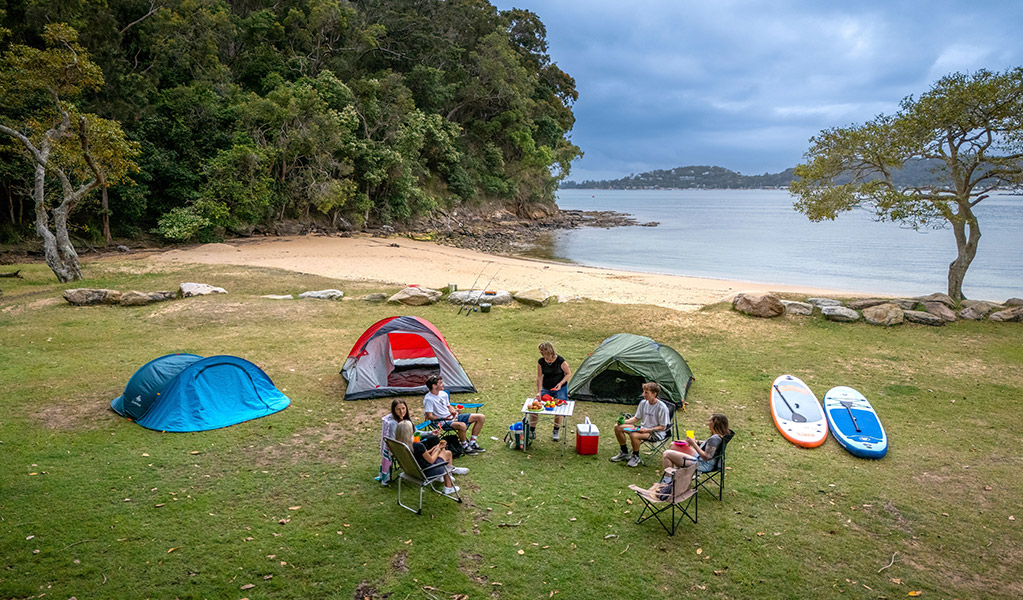 A group of campers sitting near their tents by the beach and bush at The Basin campground. Credit: John Spencer &copy; DCCEEW