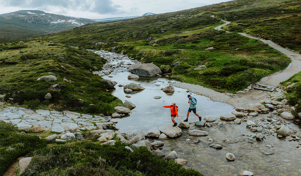 An aerial image of 2 people crossing the Snowy River by rock hopping along Snowies Alpine Walk. Credit: Daniel Parsons / DCCEEW &copy; Caravel Content