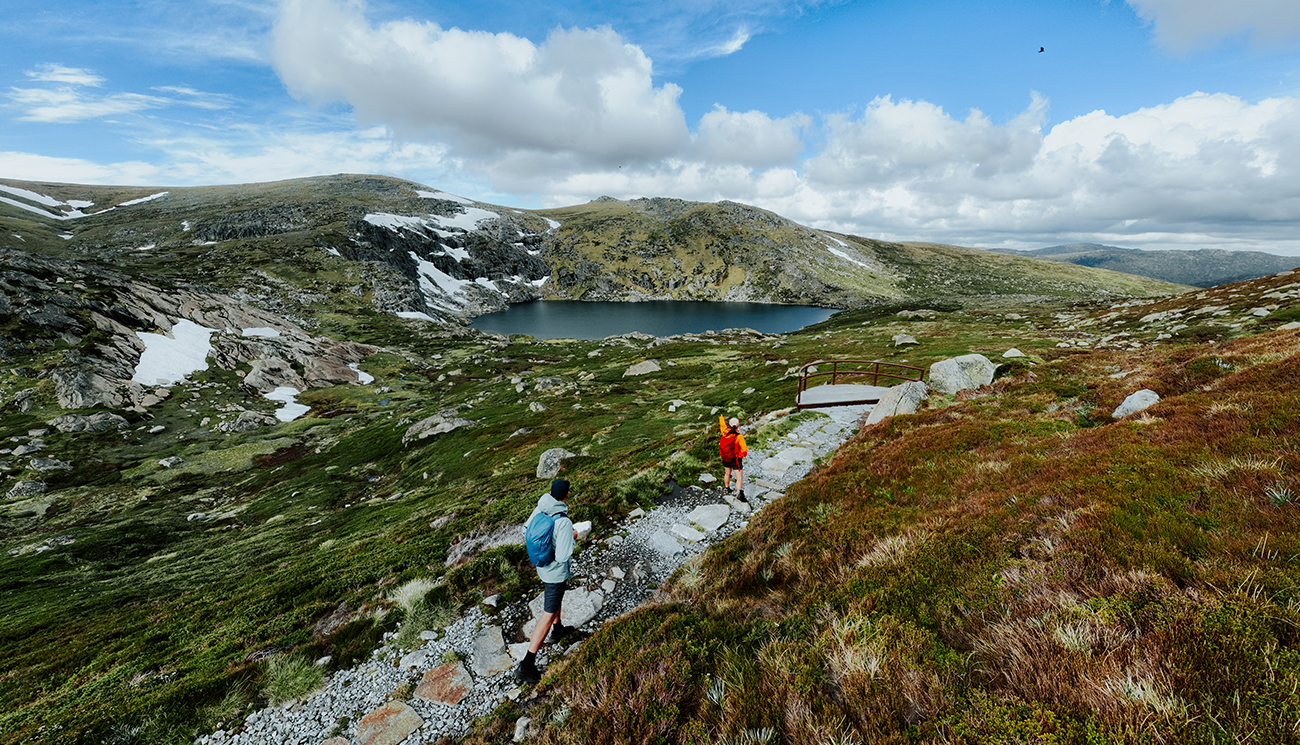 2 hikers walking on a gravel path on day 2 of Snowies Alpine Walk with Blue Lake and snow capped mountains in the background. Credit: Daniel Parsons / DCCEEW &copy; Caravel Content