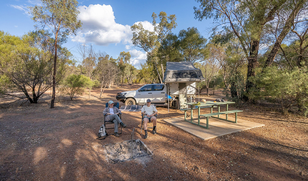 Two people sitting near picnic tables, a campfire and their tent and car at Woolshed Flat campground in Cocoparra National Park. Credit: Jonh Spencer &copy; DCCEEW