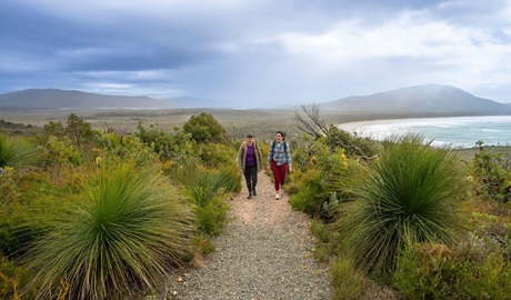 2 people on a track along Diamond Head loop walk surrounded by coastal heath with Dunbogan beach and Perpendicular Point in the background. Credit: John Spencer &copy; DCCEEW
