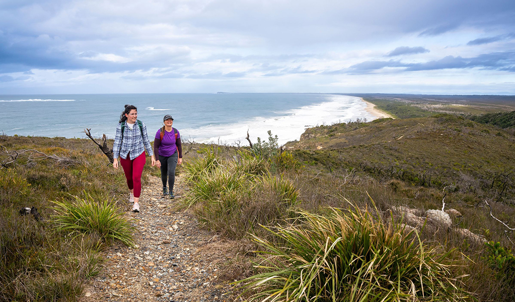 2 people bushwalking along Diamond Head loop walk in Crowdy Bay National Park with Kylies beach in the background. Credit: John Spencer &copy; DCCEEW