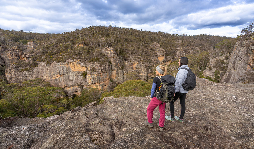 2 people looking out at a view of dramatic cliffs and thick bushland along Broad Swamp to Birds Rock walking track in Gardens of Stone State Conservation Area. Credit: John Spencer &copy; DCCEEW