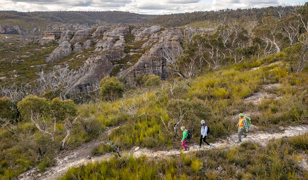3 people walking Broad Swamp to Birds Rock walking track with sweeping views of pagoda rock formations in the background. Credit: John Spencer &copy; DCCEEW