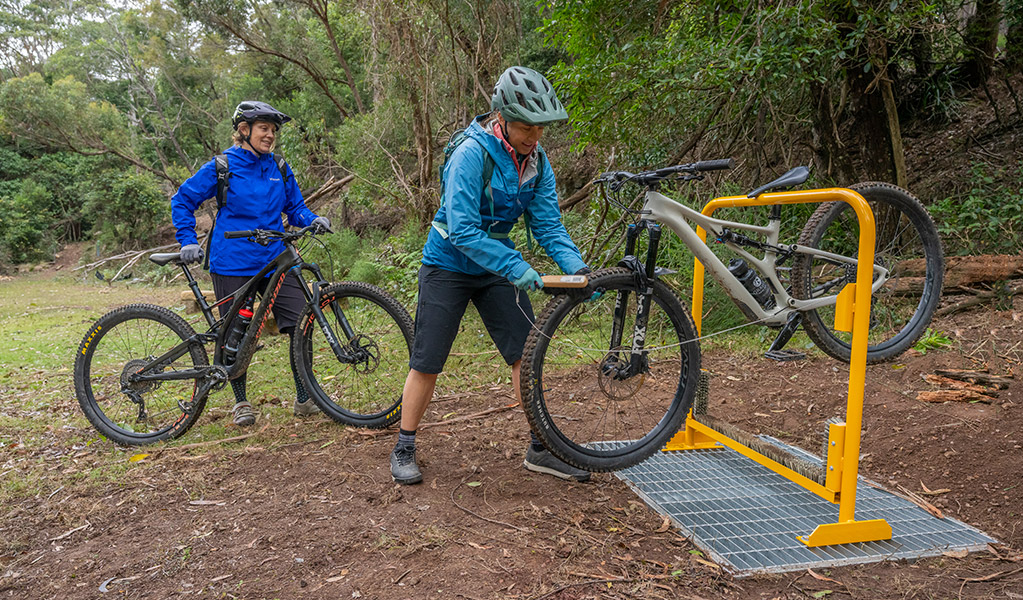 Two cyclists brush down their bikes at a cleaning station to prevent the spread of weeds. Credit: John Spencer &copy; DCCEEW