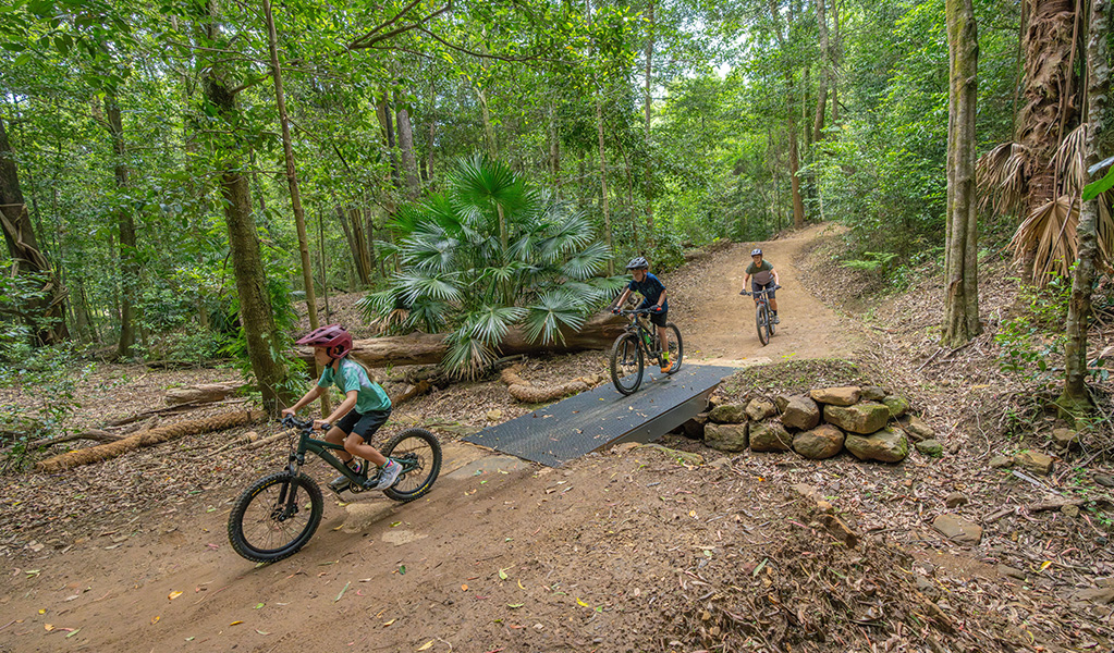 3 young kids on bikes cross a mesh bridge on a flat section of Kembla mountain bike trails set in tall forest. Credit: John Spencer &copy; DCCEEW