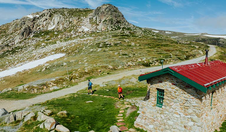 2 hikers on Main Range walk with historic Seamans Hut in the foreground and snow capped mountains in the background. Credit: Daniel Parsons / DCCEEW &copy; Caravel Content