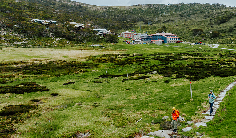 2 people walking on a stepping stone path surrounded by alpine grassland with Charlotte Pass Village in the background. Credit: Daniel Parsons / DCCEEW &copy; Caravel Content
