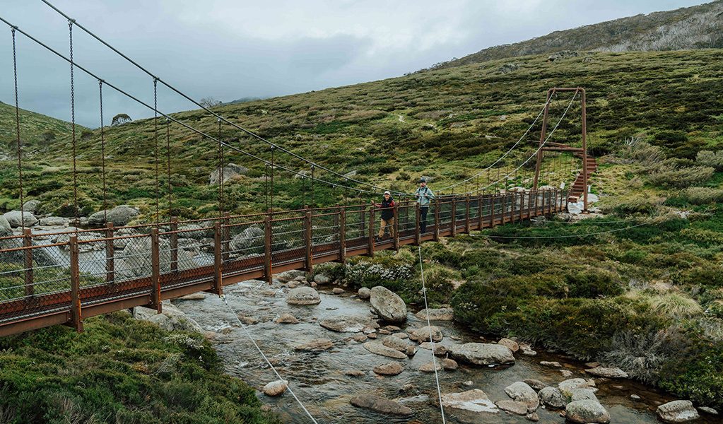 2 people crossing Spencers Creek suspension bridge with a flowing creek and alpine landscape in the background. Credit: Remy Brand / DCCEEW &copy; Caravel Content