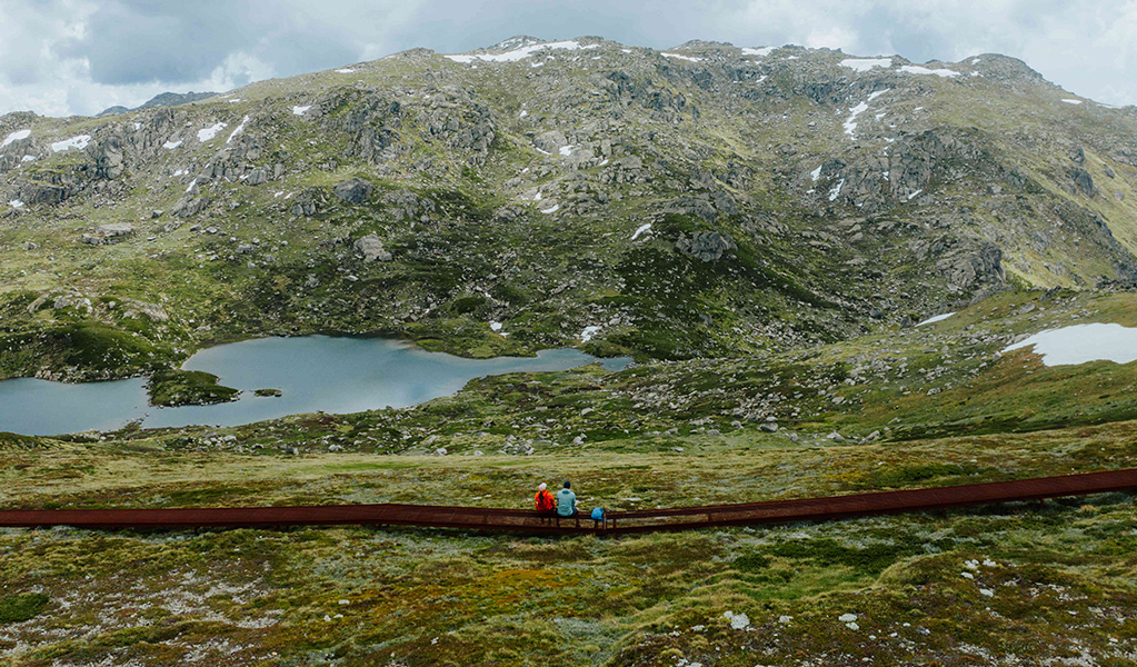 2 hikers on a raised path admiring snow capped Main Range mountain views on day 2 of Snowies Alpine Walk. Credit: Daniel Parsons / DCCEEW &copy; Caravel Content