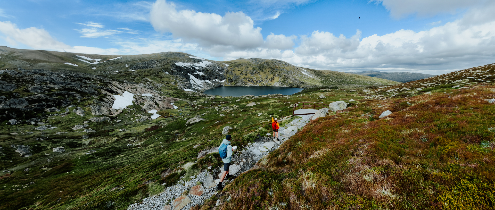 2 hikers walking Snowies Alpine Walk with a view of Blue Lake and snow capped mountain peaks in the background. Credit: Daniel Parsons / DCCEEW &copy; Caravel Content