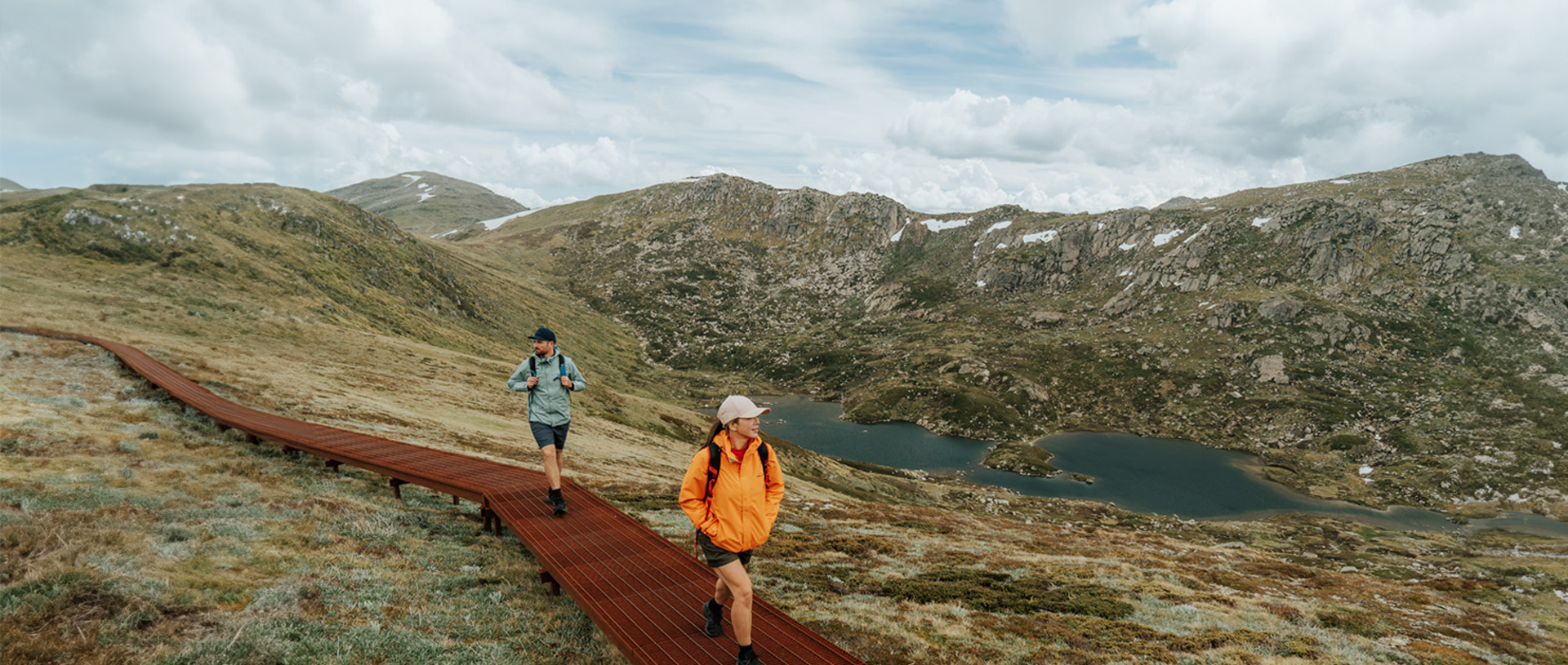 2 hikers walking on a raised path on day 2 of Snowies Alpine Walk with snow capped mountains in the background. Credit: Remy Brand / DCCEEW &copy; Caravel Content