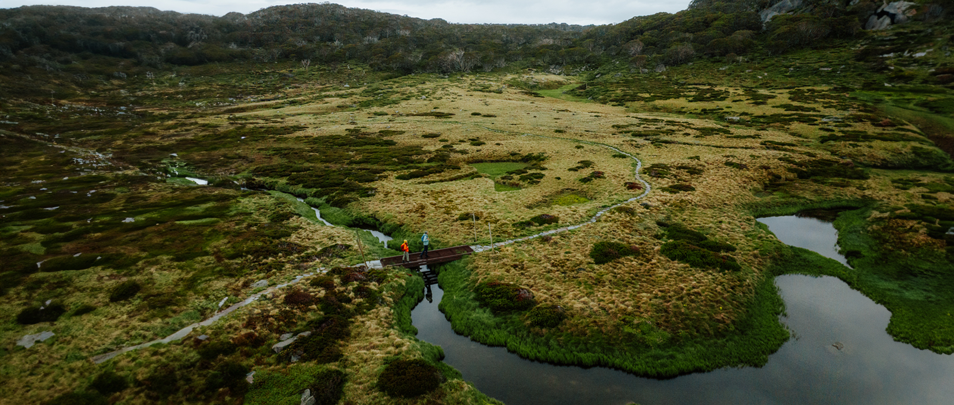 An aerial view of 2 hikers crossing a winding creek surrounded by marsh on Snowies Alpine Walk, Kosciuszko National Park. Credit: Daniel Parsons / DCCEEW &copy; Caravel Content