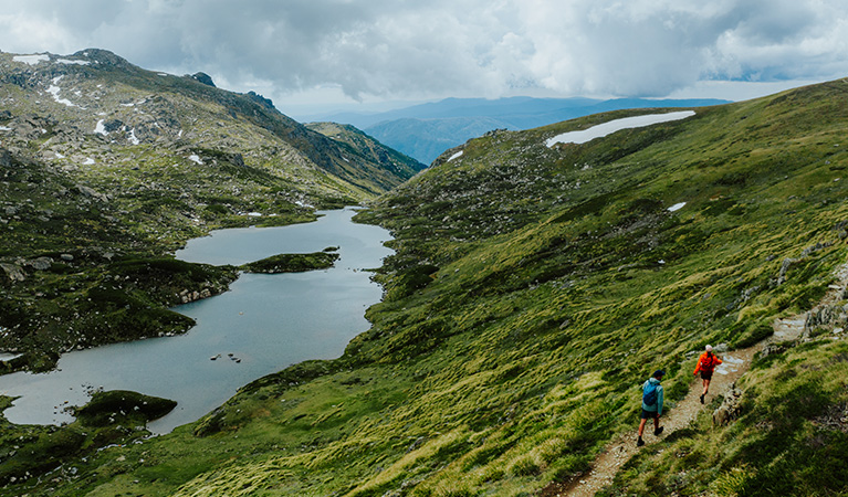 2 hikers on a track along Snowies Alpine Walk with a view of snow-capped mountains and a lake in the background. Credit: Daniel Parsons / DCCEEW &copy; Caravel Content