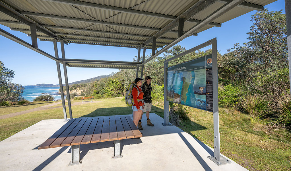 Visitors reading a park sign under shelter at Pretty Beach picnic area. Credit: John Spencer &copy; DCCEEW