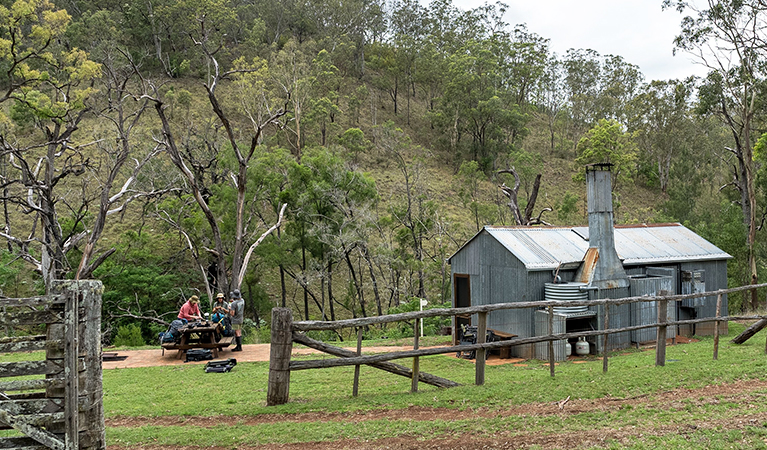Corrugated iron Green Gully Hut located next to a creek with walkers seated at an outdoor picnic table. Credit: David Waugh &copy; DCCEEW