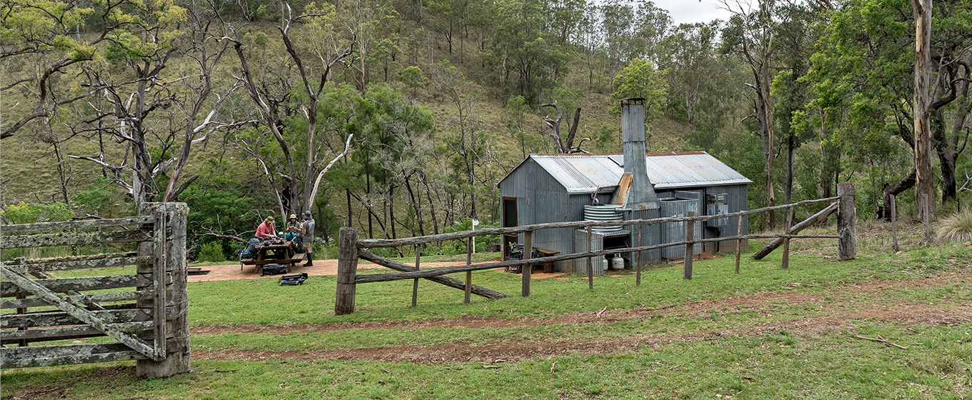 A group of multi-day hikers sitting at a picnic table beside the rustic Green Gully Hut surrounded by trees. &copy; DCCEEW