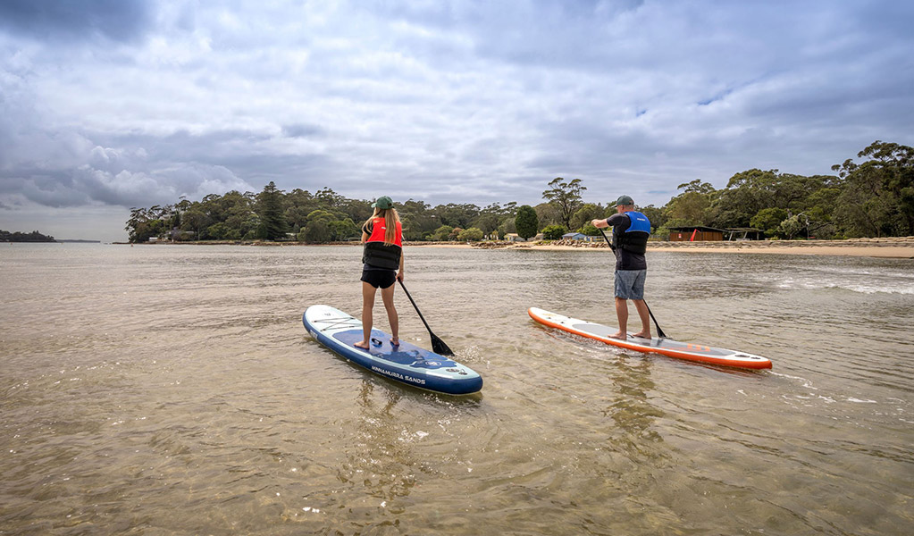 Two people paddle boarding on Hacking river next to Bonnie Vale campground. Credit: John Spencer &copy; DCCEEW