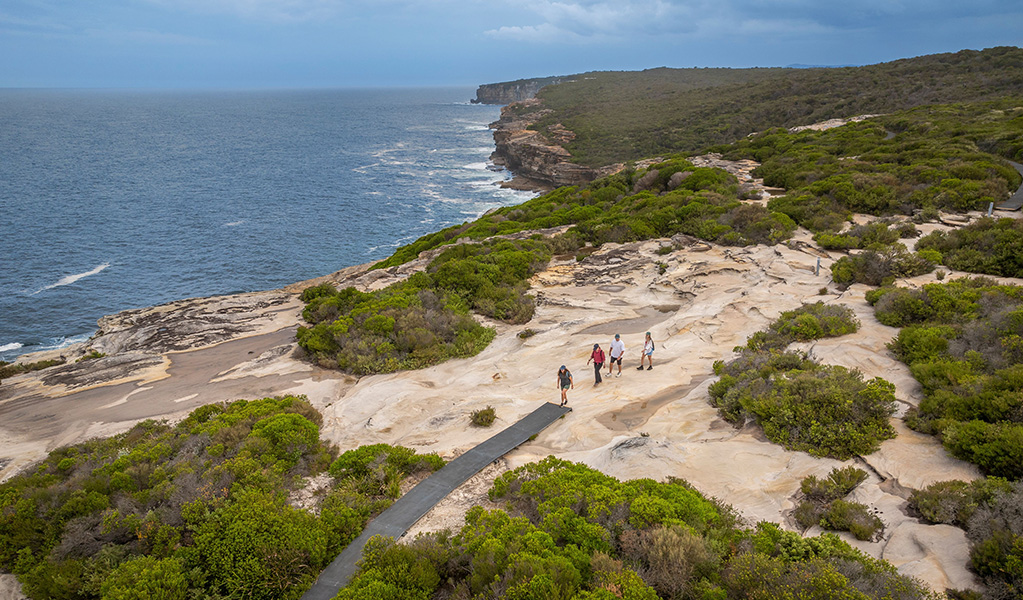 A group of hikers on The Coast Track with the sandstone cliffs and the ocean in the background. Credit: John Spencer / DCCEEW &copy; DCCEEW