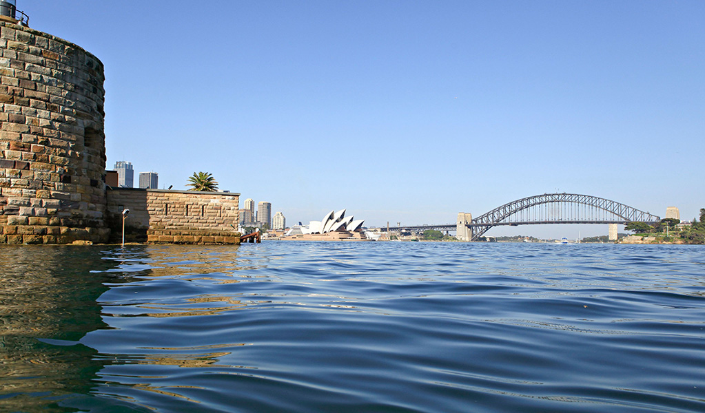 View across Sydney Harbour of Fort Denison's round sandstone Martello Tower with the Harbour Bridge and Opera House in the background. Credit: Rosie Nicolai &copy; DCCEEW