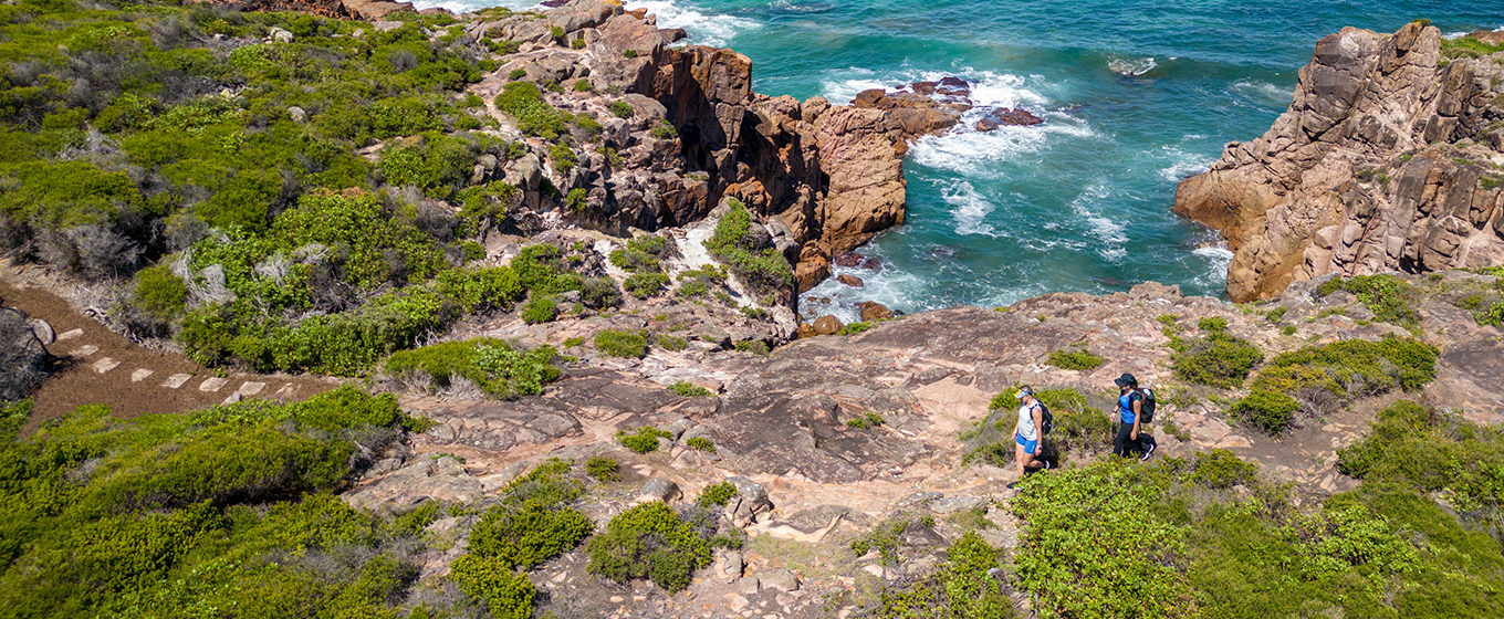 Aerial view looking down at two walkers approaching stepping stones on the edge of coastal rock platform. Credit: John Spencer &copy; DPE