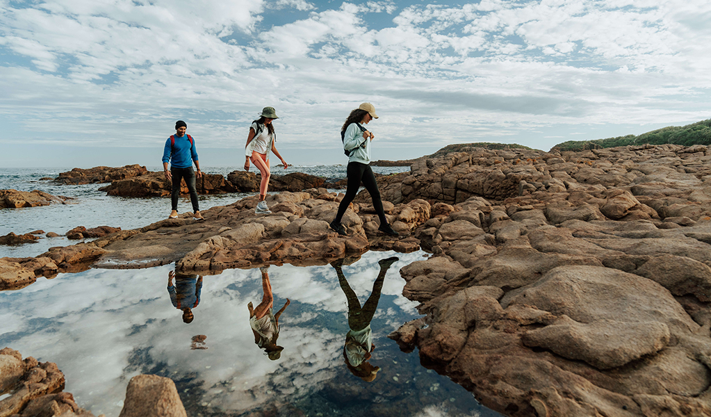 Walkers crossing a rock platform with calm ocean rock pool casting their reflection along Tomeree Coastal Walk. Credit: Remy Brand &copy; Remy Brand
