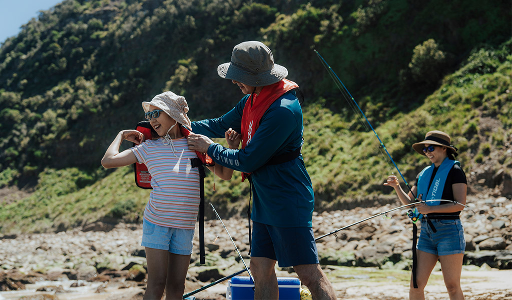 A person helps another person to put on a rock fishing lifejacket when fishing off the rocks in a national park. Credit: Remy Brand/DCCEEW